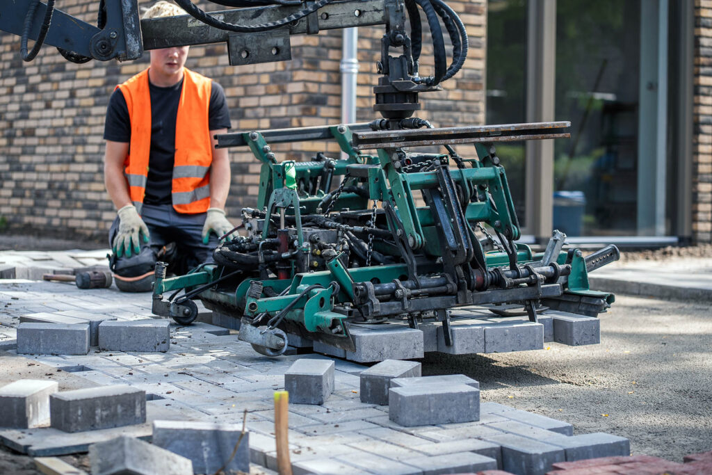 Bestratingsklem plaatst grijze betonklinkers op een zandbed, begeleid door een stratenmaker naast een woning.