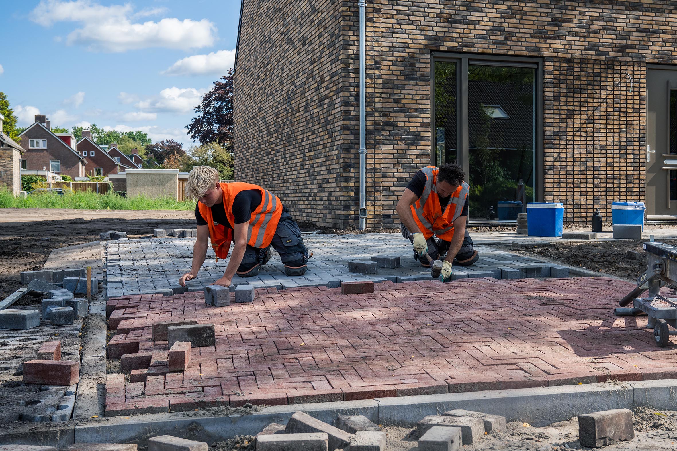Aanleg van bestrating bij een woning door Henk Verhoef BV. Twee vakmannen leggen grijze klinkers en werken de aansluiting op de rode bestrating zorgvuldig af.