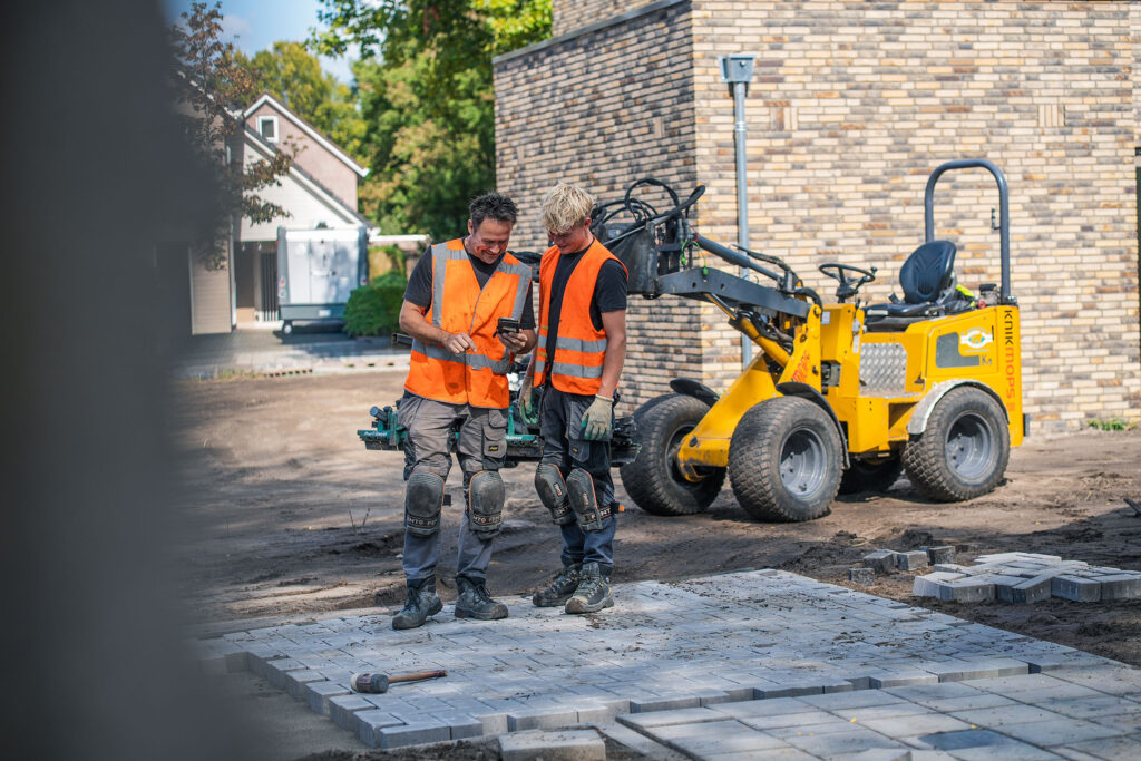 Twee vakmannen van Henk Verhoef BV bekijken samen een telefoon tijdens straatwerk, naast een gele shovel en nieuw gelegde bestrating.